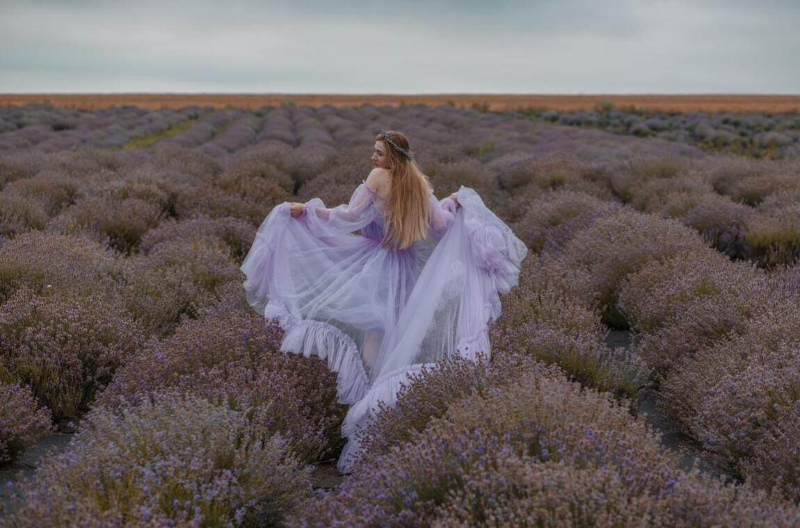 Дівчина в капелюсі та білій сукні позує серед рядів лаванди, вигляд зі спини